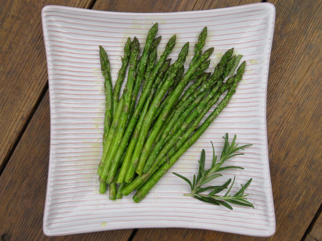 Oven roasted asparagus infused with rosemary olive oil, on a white serving plate with a fresh sprig of rosemary off to the side. 