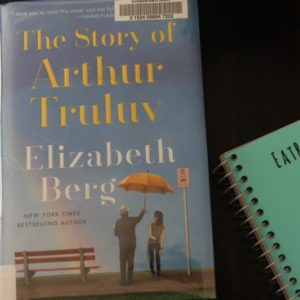 The book The Story of Arthur Truluv by Elizabeth Berg, on a wood table with the EatReadCruise notebook off to the side