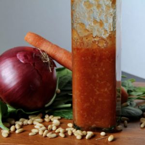 Carolyn's Creation salad dressing in a tall glass bottle with red onion, pine nuts, carrot and spinach around the dressing