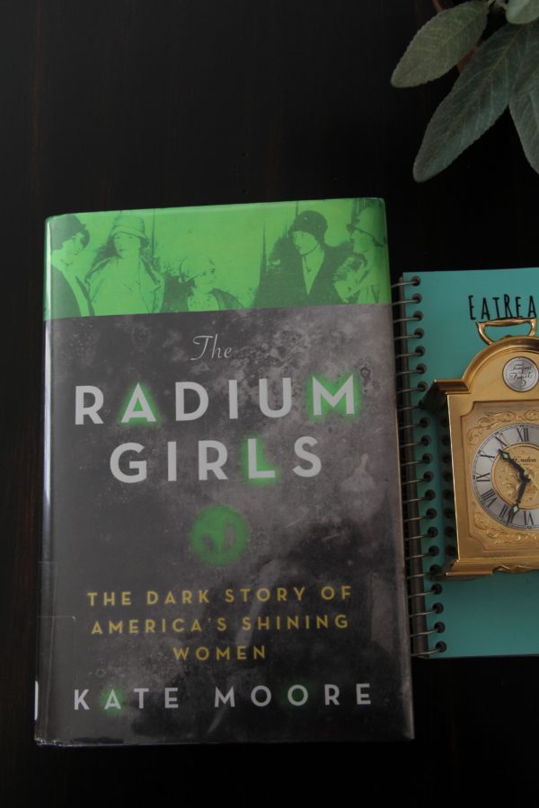 The book Radium Girls, with an EatReadCruise notebook nearby and a luminous clock on the notebook