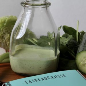 Greeg Goddess Salad Dressing in a clear glass jar with lettuce and a cumber behind it. The tea EatReadCRuise notebook is peaking on the bottom of the photo