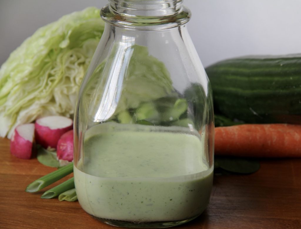 Green Goddess Salad Dressing in a clear jar with lettuse, carrots, green onions and a cucumber in the background. On a wood table.