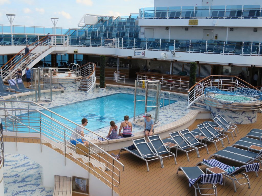Some passengers around the main pool on a Royal Princess cruise. Included are two hot tubs, each ona different side of the pool.