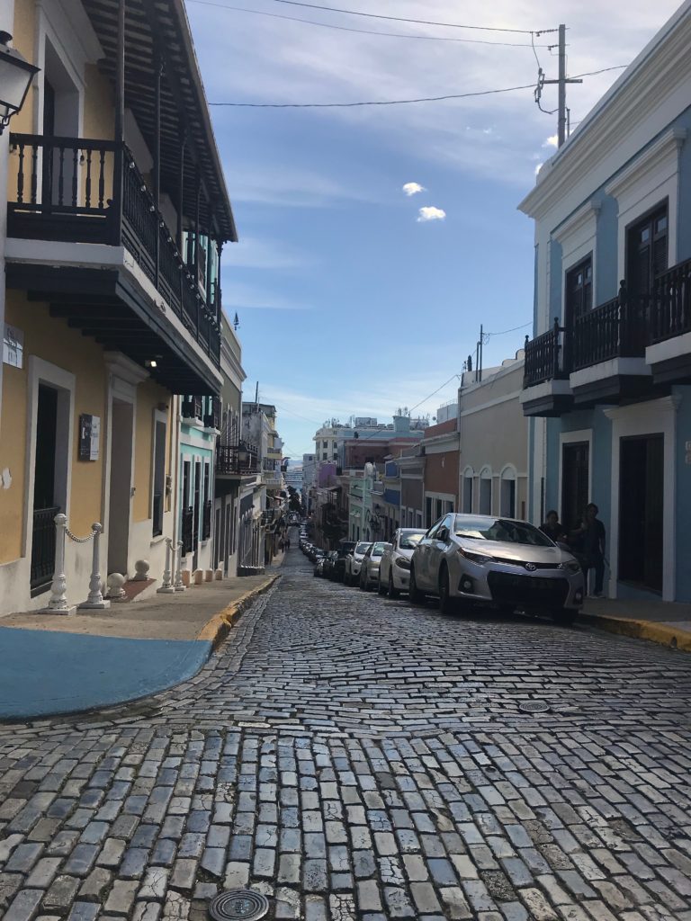 Narrow, cobblestone street in Old San Juan, near town with cars parked along the right side of the street