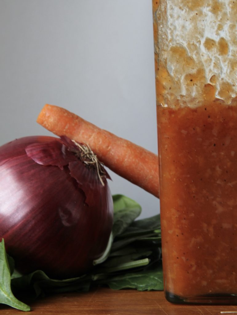 Carolyn's salad dressing, in a tall clear glass bottle, red in color, on a wood table with a red onion, orange carrot and spinach in the background, ready to be made into a tossed green salad