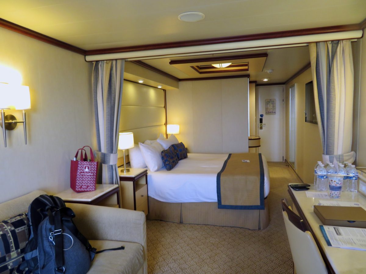 Photo of a mini-suite on the Regal Princess taken from the balcony. Shows the couch, desk, queen bed and 2 nightstands. 