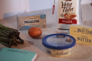 Ingredients for the asparagus appetizer including a loaf of thin slice white bread, 2 sticks of butter, fresh asparagus, 4 oz. container of crumbled blue cheese, an egg and 8 oz. of cream cheese, with the teal EatReadCruise notebook peaking in the left bottom corner.
