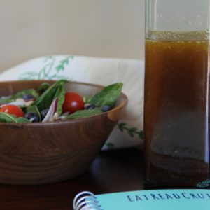 Teriyaki salad dressing, in a tall clear glass bottle, to the left of an individual green salad in a wood bowl. The teal EatReadCruise notebook is peaking below.