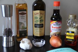 Ingredients for the teriyaki salad dressing on a wood table including Teriyaki sauce, white wine vinegar, fesh garlic, fresh onion, salt, pepper, honey and extra virgin olive oil.