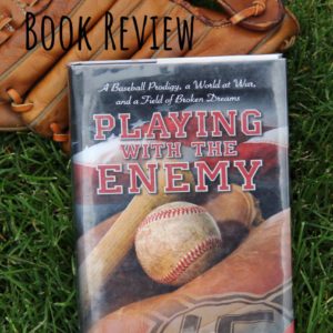 The hardcover book, Playing With the Enemy, lying on green grass, propped up on a leather baseball glove.