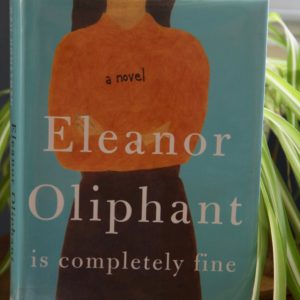 Hardbook copy of Eleanoe Oliphant is ompletely Fine on a wood table, in fromt of window with blinds and a green spider plant on the side of book, book review, book club questions,