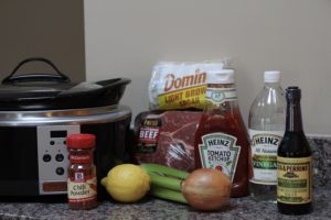 Ingredients for barbecue beef including beef roast, brown sugar, vinegar, chili powder, lemon, onion, celery and catsup. Slow cooker on side of ingredients