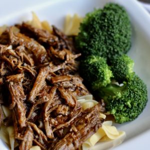 Berbecue beef, shredded, on wide egg noodles, on a white plate along with steamed broccoli