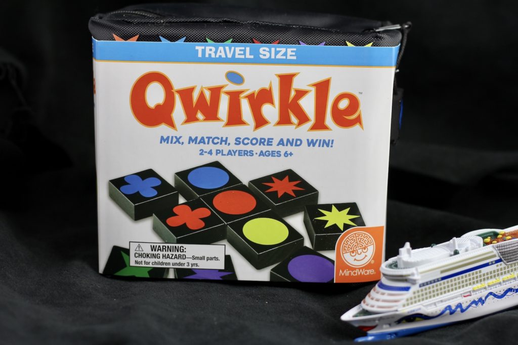 Tile travel game Qwirkle, on black background, with a small model of a cruise ship peaking in lower right corner of photo