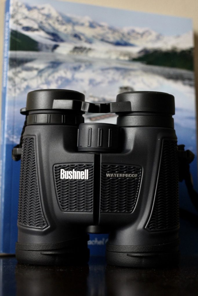 Black Bushnell waterproof binoculars in from of an Alaska book. Alaska cruise tips