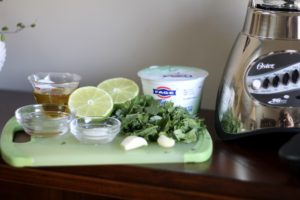 Ingredients for cilantro salad dressing sitting next to an electric blender including cilantro, fresh garlic, salt, lime juice, Greek yogurt, white vinegar and olive oil.